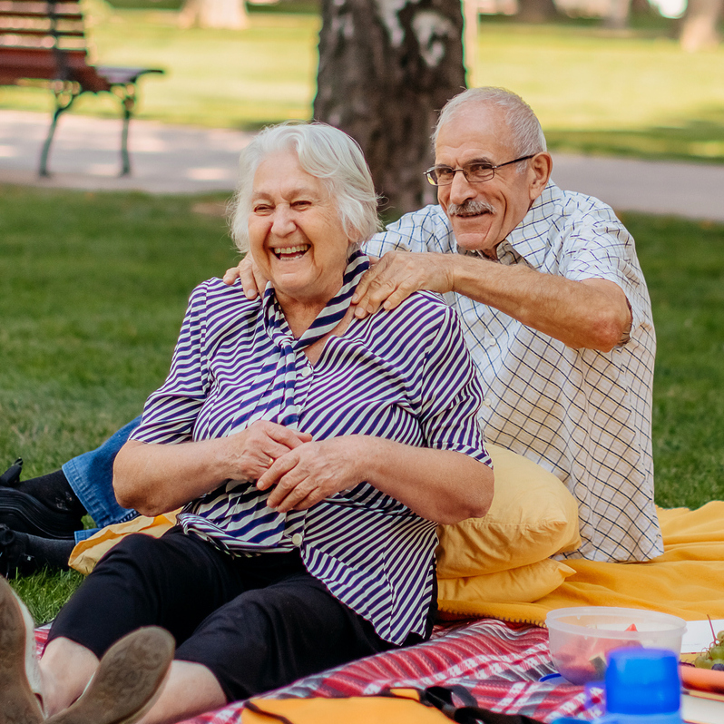 Auf dem Foto sieht man eine ältere Frau und einen älteren Mann. Sie sitzen auf einer Picknickdecke im Park und massieren sich. 